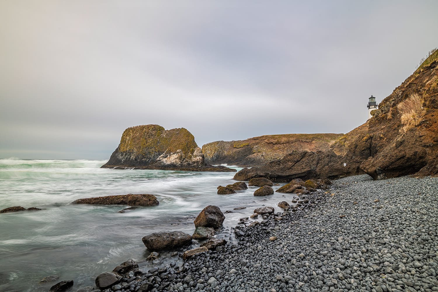 Long-exposure surf smoothing to mist around basalt sea stacks and a cobble beach, with a small lighthouse standing on the bluff above, Oregon Coast, in flat evening light.