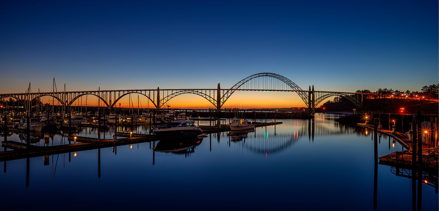 The lit steel arches of the Yaquina Bay Bridge spanning the marina at Newport, Oregon, at blue hour, with a band of orange sunset still on the horizon and the moored boats and dock lights mirrored in flat water.